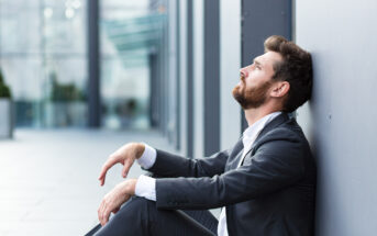 A man in a suit sits on the ground, leaning against a wall, looking upwards in an urban setting. The background is blurred, suggesting a modern building exterior.