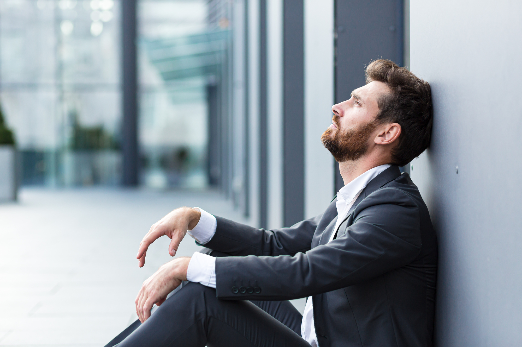 A man in a suit sits on the ground, leaning against a wall, looking upwards in an urban setting. The background is blurred, suggesting a modern building exterior.