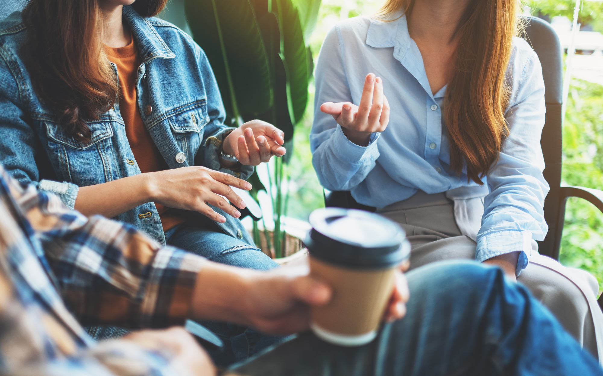 Three people are sitting and engaged in conversation. One has a denim jacket, another a blue shirt, and one is wearing plaid. A coffee cup is held by one person. Green plants are visible in the background.