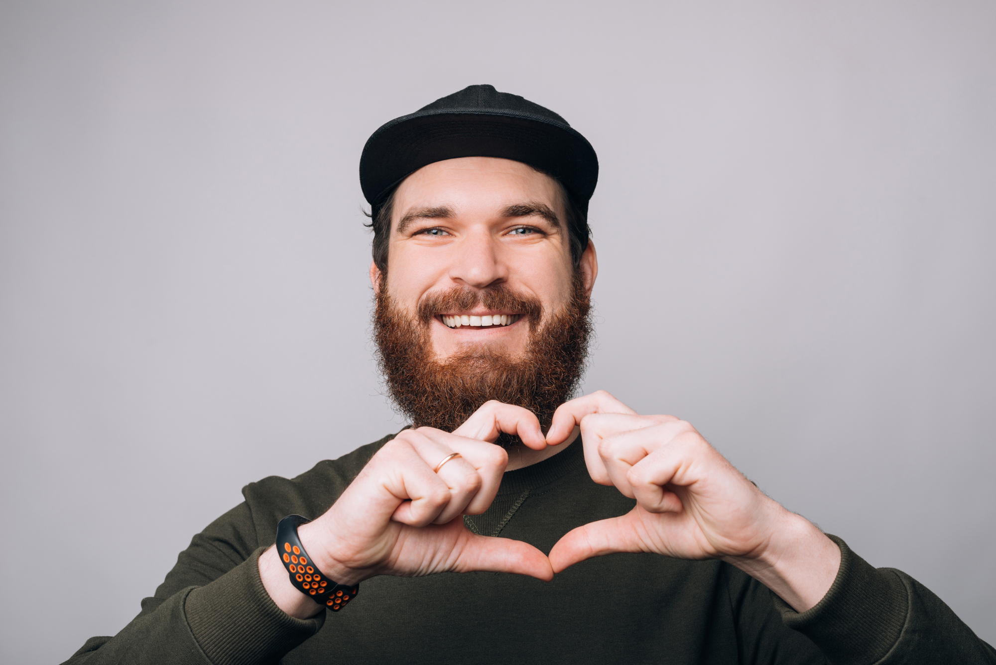 A bearded man wearing a dark cap and a green sweater forms a heart shape with his hands, smiling against a plain gray background.