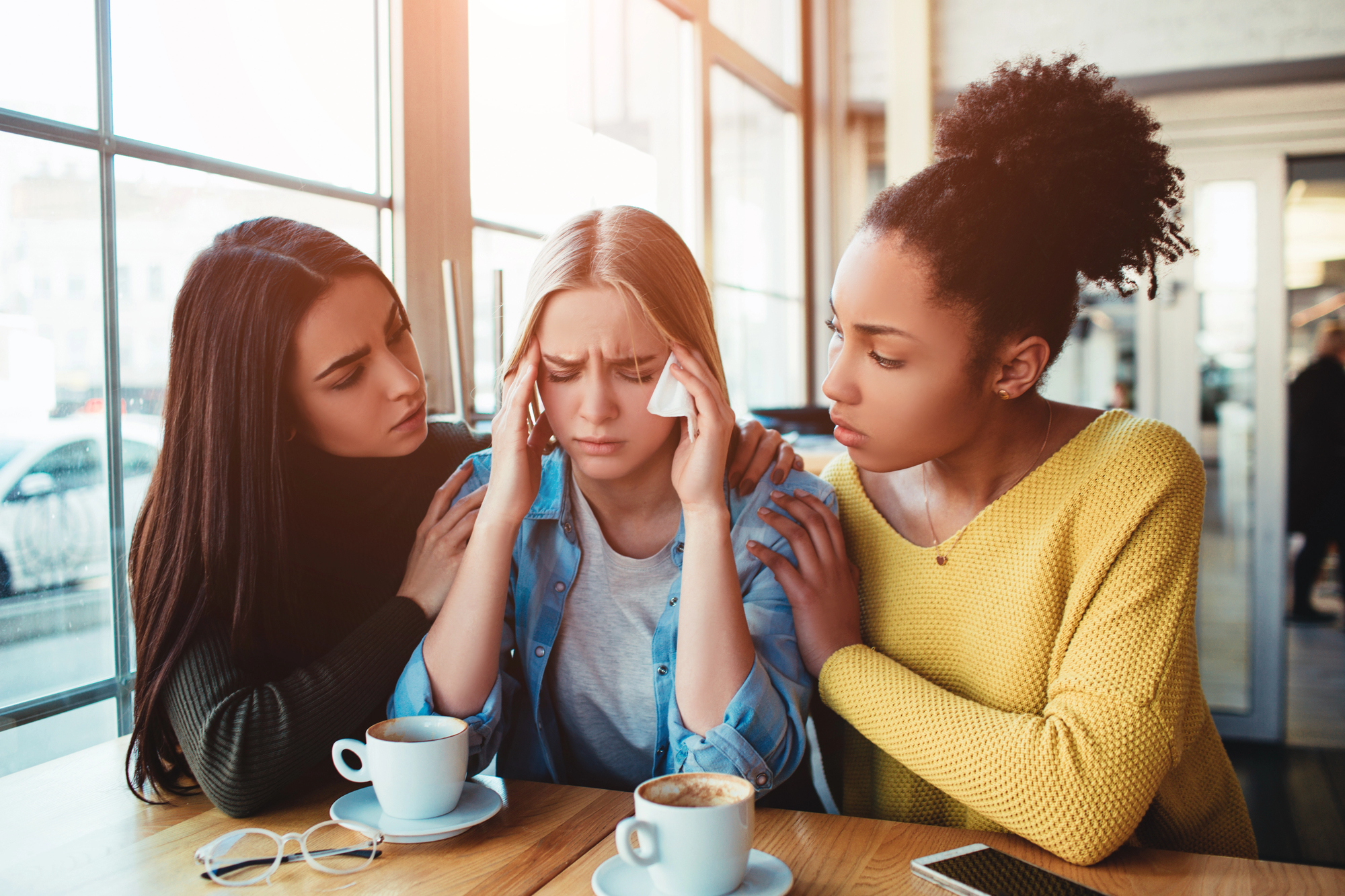 Three women sitting at a table in a café. The woman in the middle appears distressed, holding her head with her hands. The other two women are comforting her with concerned expressions. There are coffee cups on the table and sunlight coming through the window.