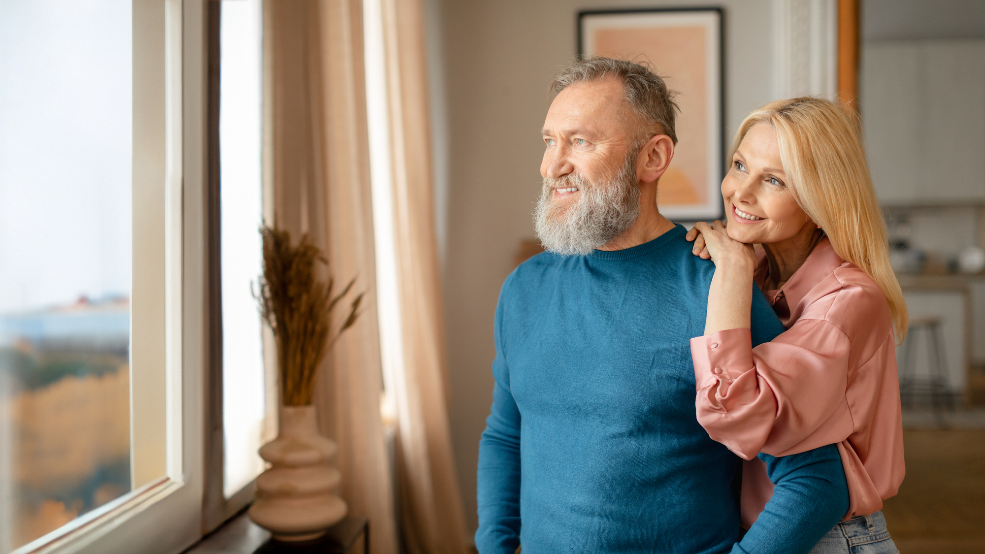 A smiling couple stands in a cozy room, gazing out a window. The man with a beard wears a blue sweater, and the woman with long blonde hair rests her hand on his shoulder, wearing a pink blouse. They appear content and relaxed.