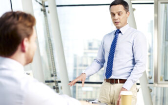 Two men are in an office with large windows. One man stands, wearing a light blue shirt and blue tie, gesturing as he talks. The other man, sitting with his back to the camera, listens attentively. A coffee cup and documents are on the desk in front of them.