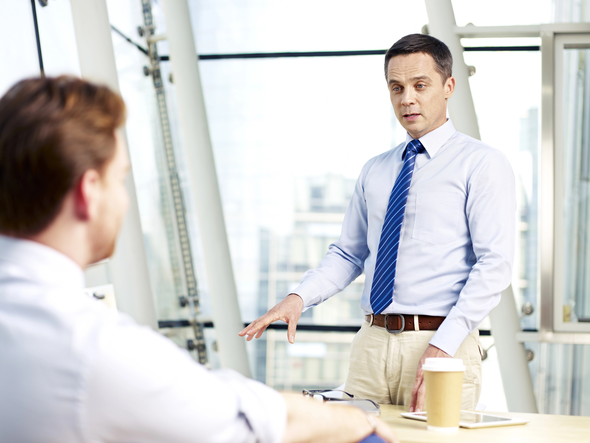 Two men are in an office with large windows. One man stands, wearing a light blue shirt and blue tie, gesturing as he talks. The other man, sitting with his back to the camera, listens attentively. A coffee cup and documents are on the desk in front of them.