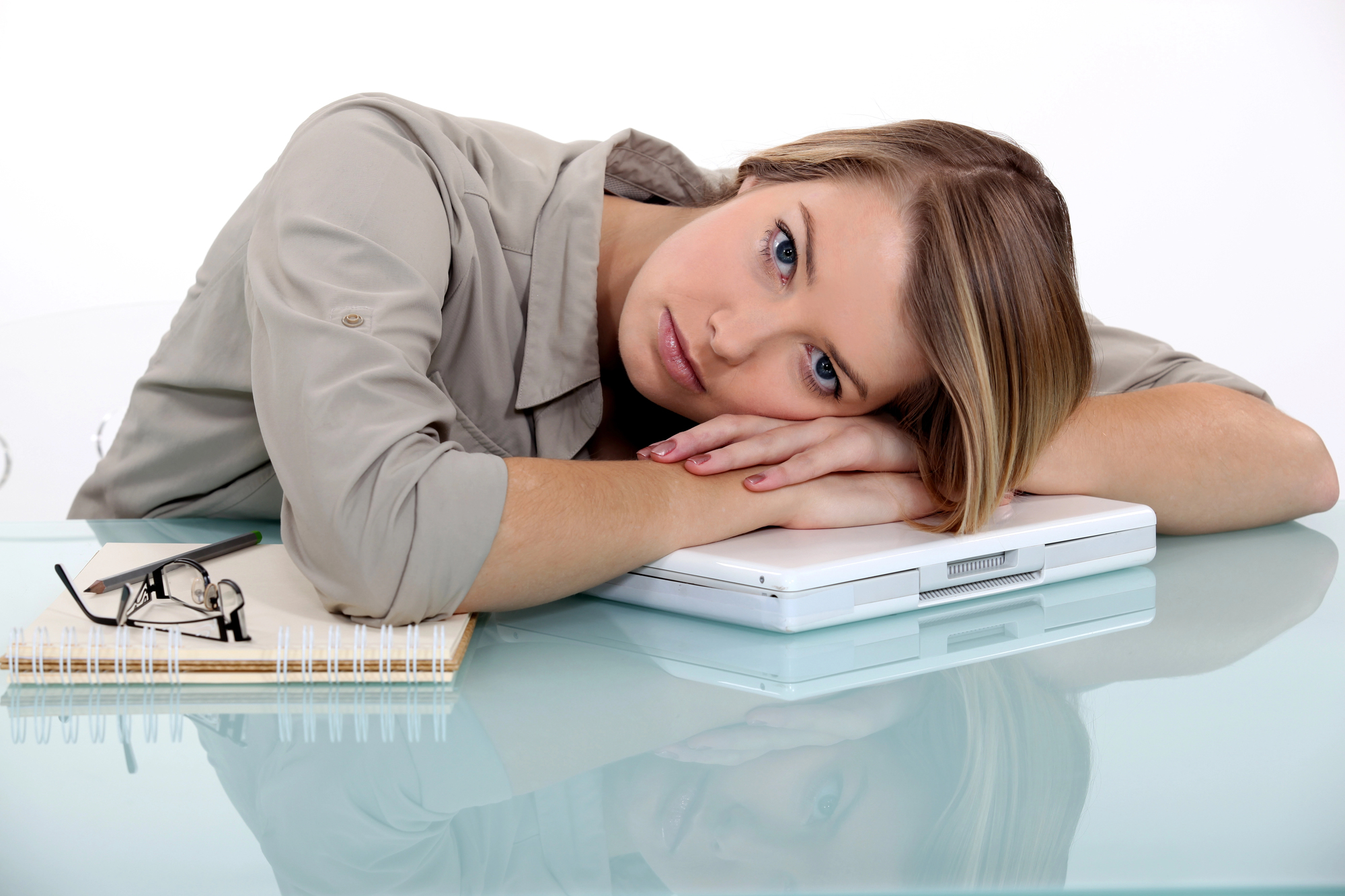 A woman rests her head on a closed laptop, looking at the camera. She is sitting at a glass desk with a notebook and glasses beside her. She appears contemplative or tired, wearing a light gray shirt.