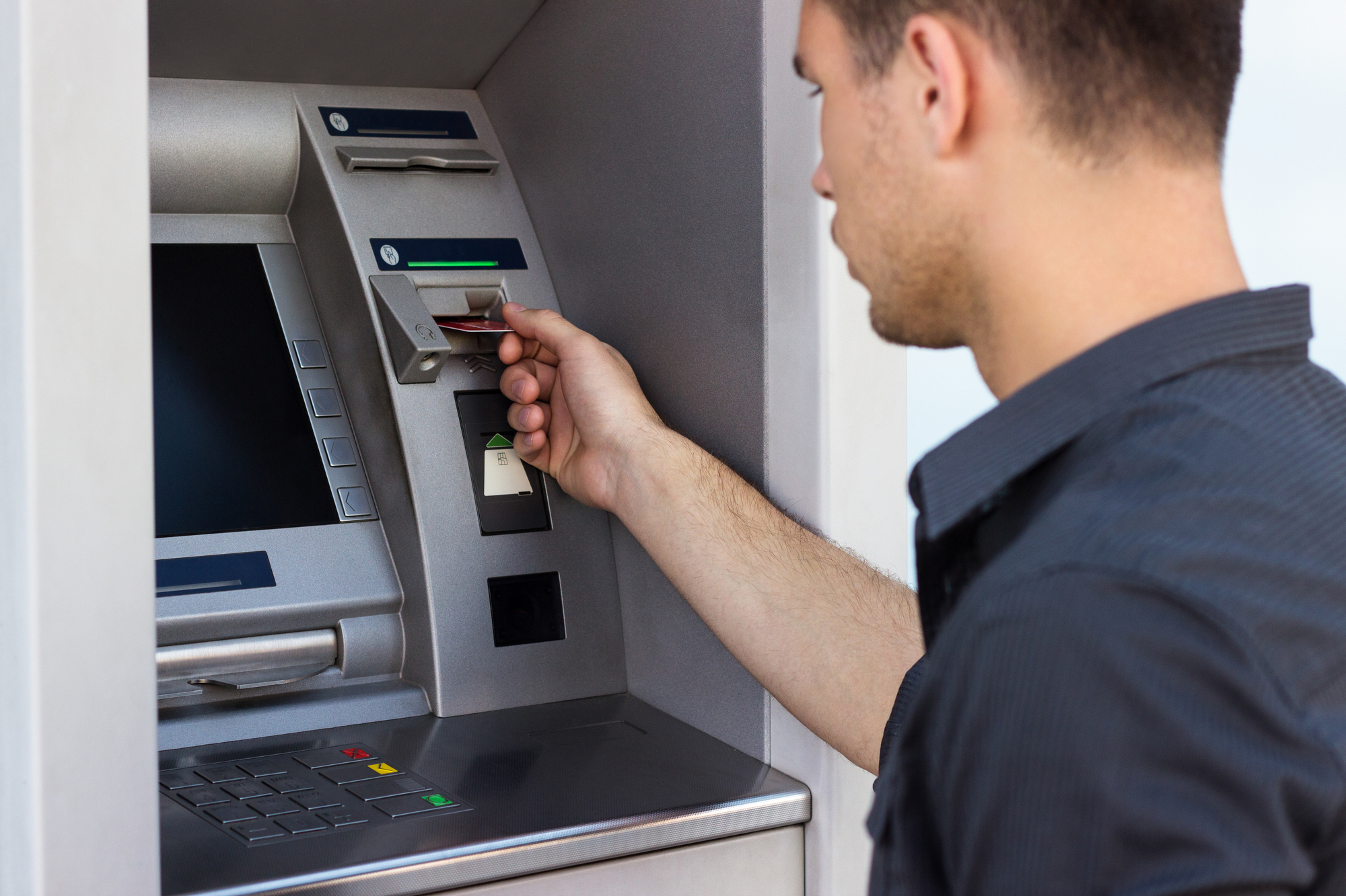 A person in a dark shirt inserts a card into an ATM machine, preparing to conduct a transaction. The ATM has a screen, keypad, and several card slots visible.