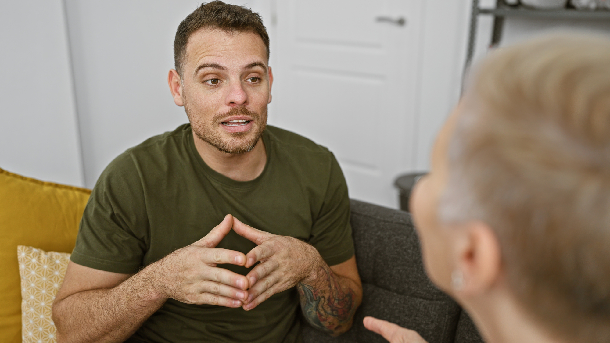 A man in a green shirt sits on a couch, gesturing with his hands while talking to another person whose back is to the camera. They appear to be having a conversation in a cozy living room setting.