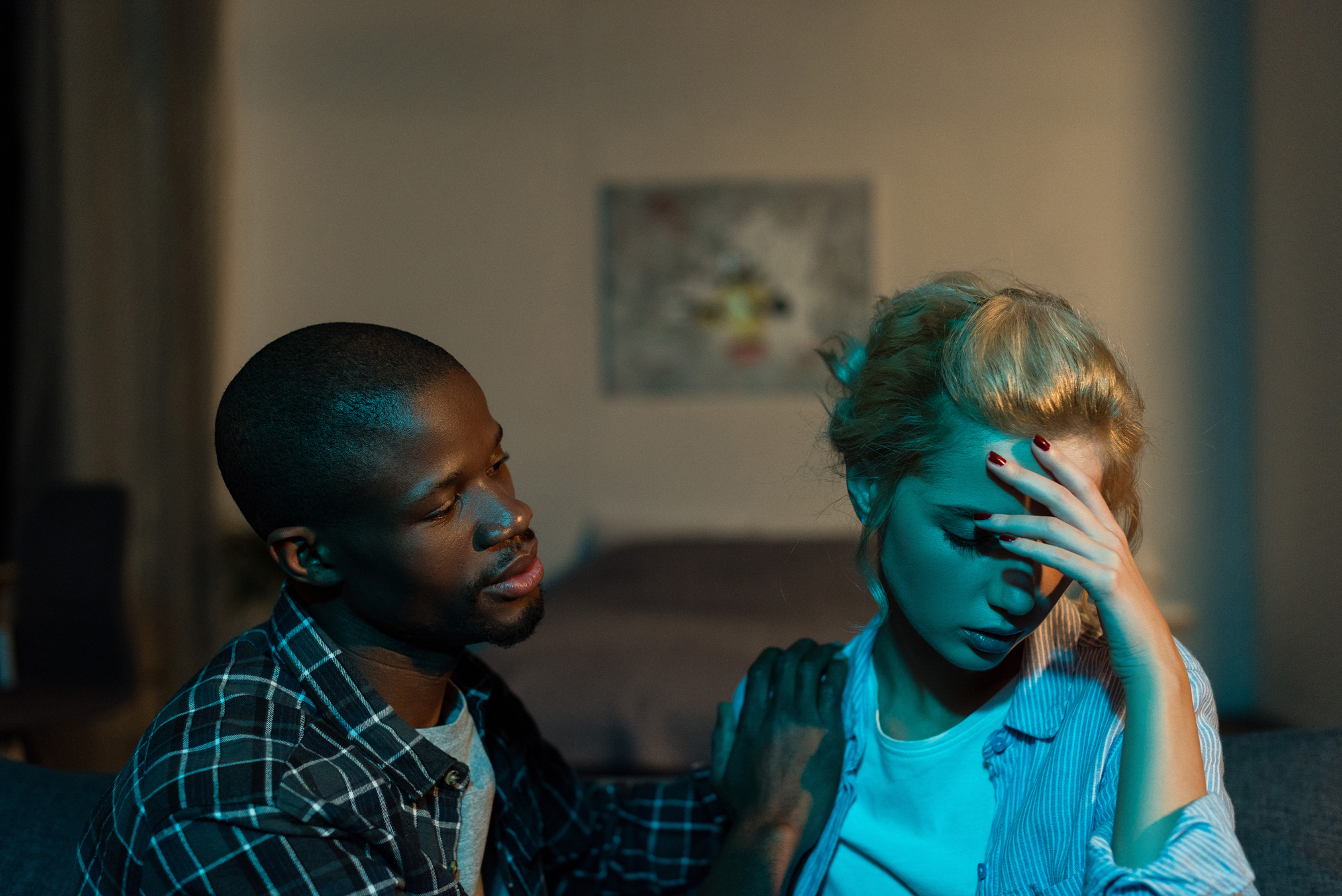 A man with a concerned expression places his hand on the shoulder of a woman who appears distressed. She is holding her head with one hand and looking down. The setting is indoors with dim lighting and a blurry painting in the background.