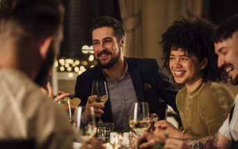 A group of people sitting around a dining table, laughing and enjoying each other's company. They have wine glasses in hand, and the atmosphere is warm and cozy, with dim lighting and a festive background.