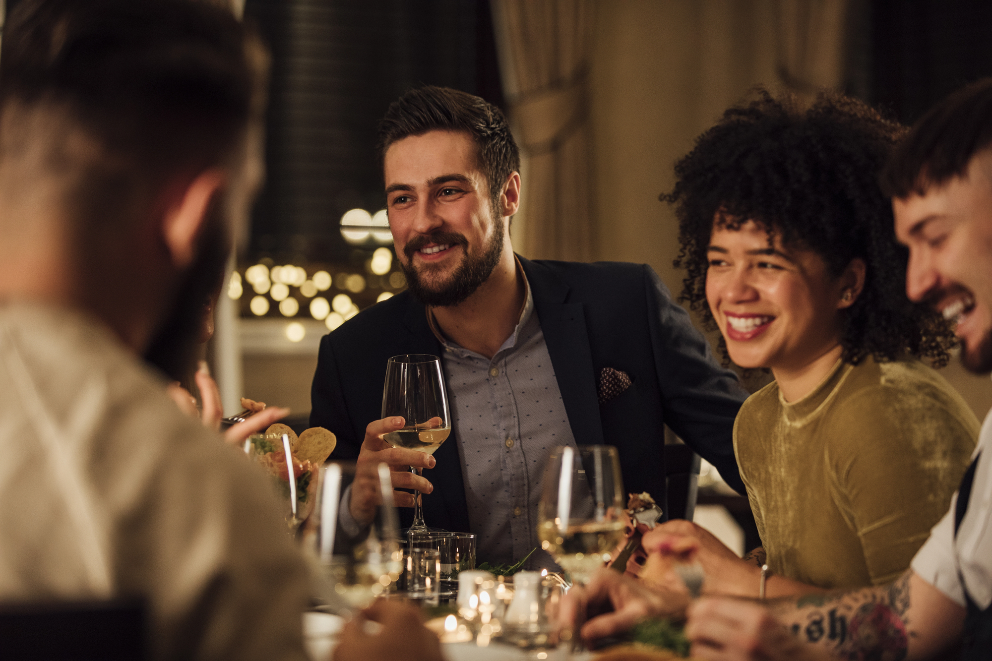 A group of people sitting around a dining table, laughing and enjoying each other's company. They have wine glasses in hand, and the atmosphere is warm and cozy, with dim lighting and a festive background.