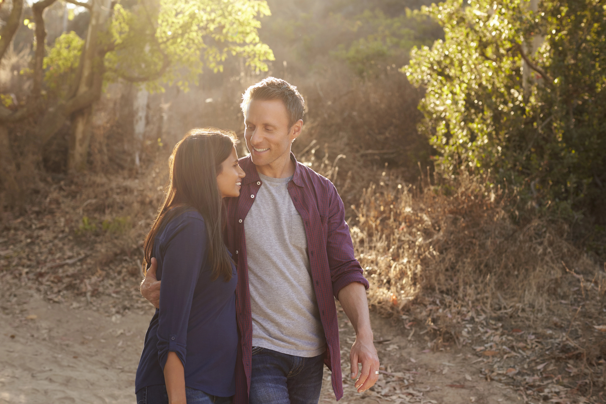 A couple walks along a sunlit path in a forest, smiling at each other. The man wears a red shirt over a gray t-shirt, and the woman wears a dark blue long-sleeve shirt. Sunlight filters through the trees, casting a warm glow.