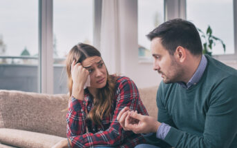 A man and a woman are sitting on a couch in a living room. The man is gesturing with his hands, explaining something, while the woman looks concerned and rests her head on her hand. Large windows are in the background.