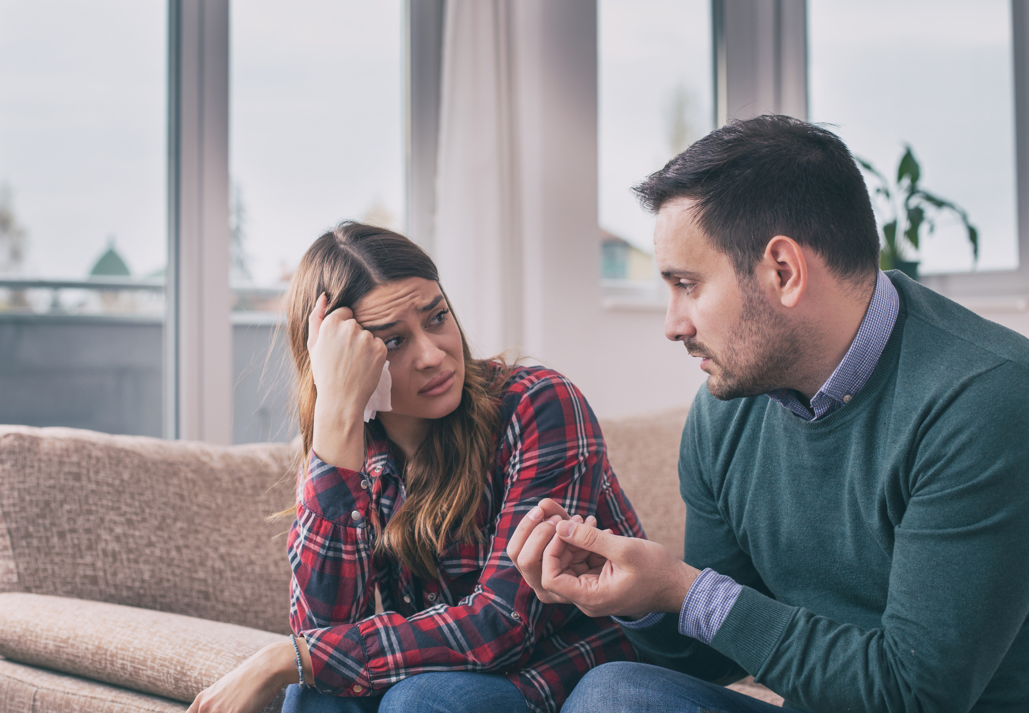 A man and a woman are sitting on a couch in a living room. The man is gesturing with his hands, explaining something, while the woman looks concerned and rests her head on her hand. Large windows are in the background.