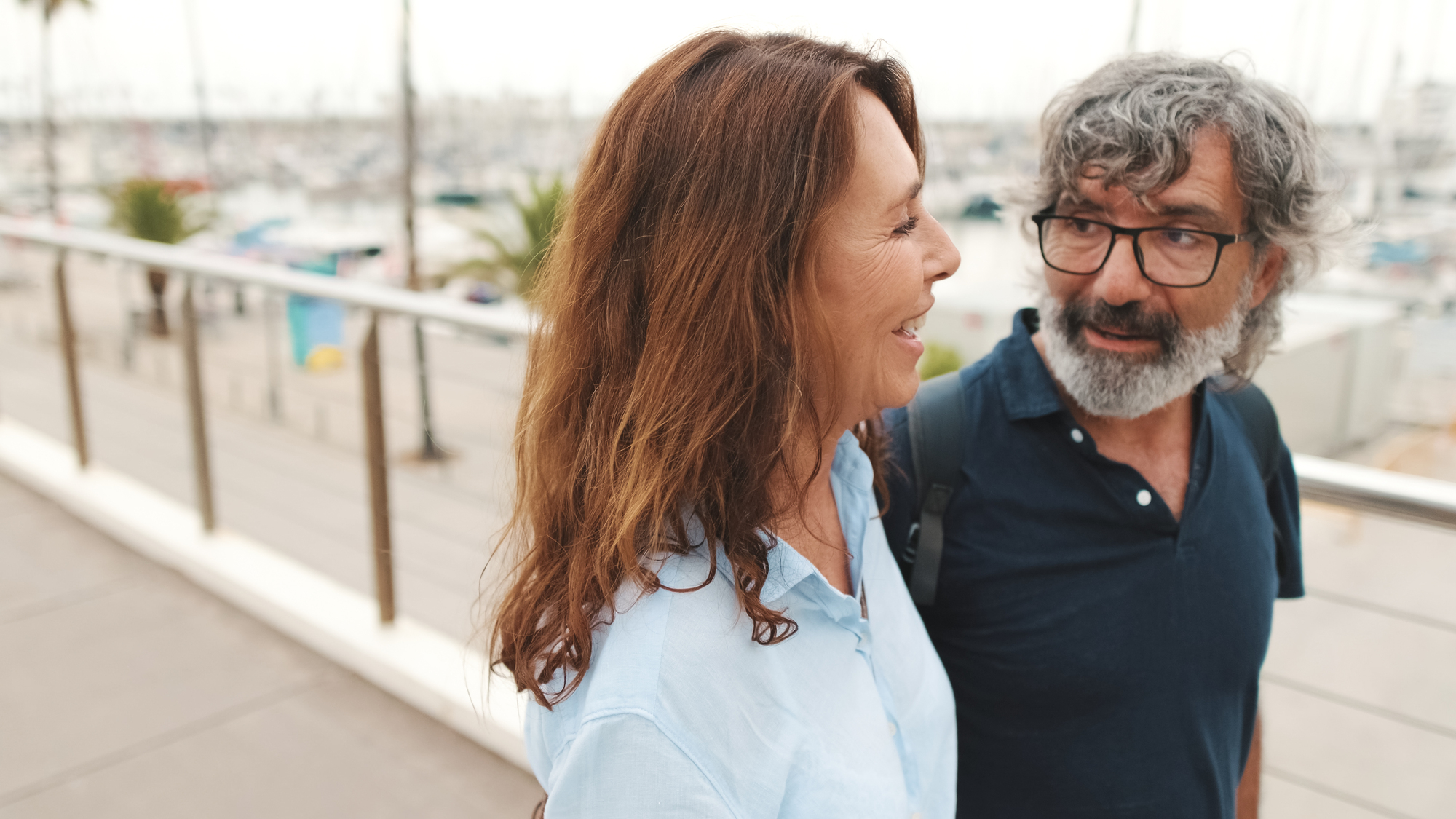 A woman and a man with gray hair walk side by side on a boardwalk overlooking a marina. Both are smiling and appear to be engaged in conversation. The background is slightly blurred, focusing on the couple.