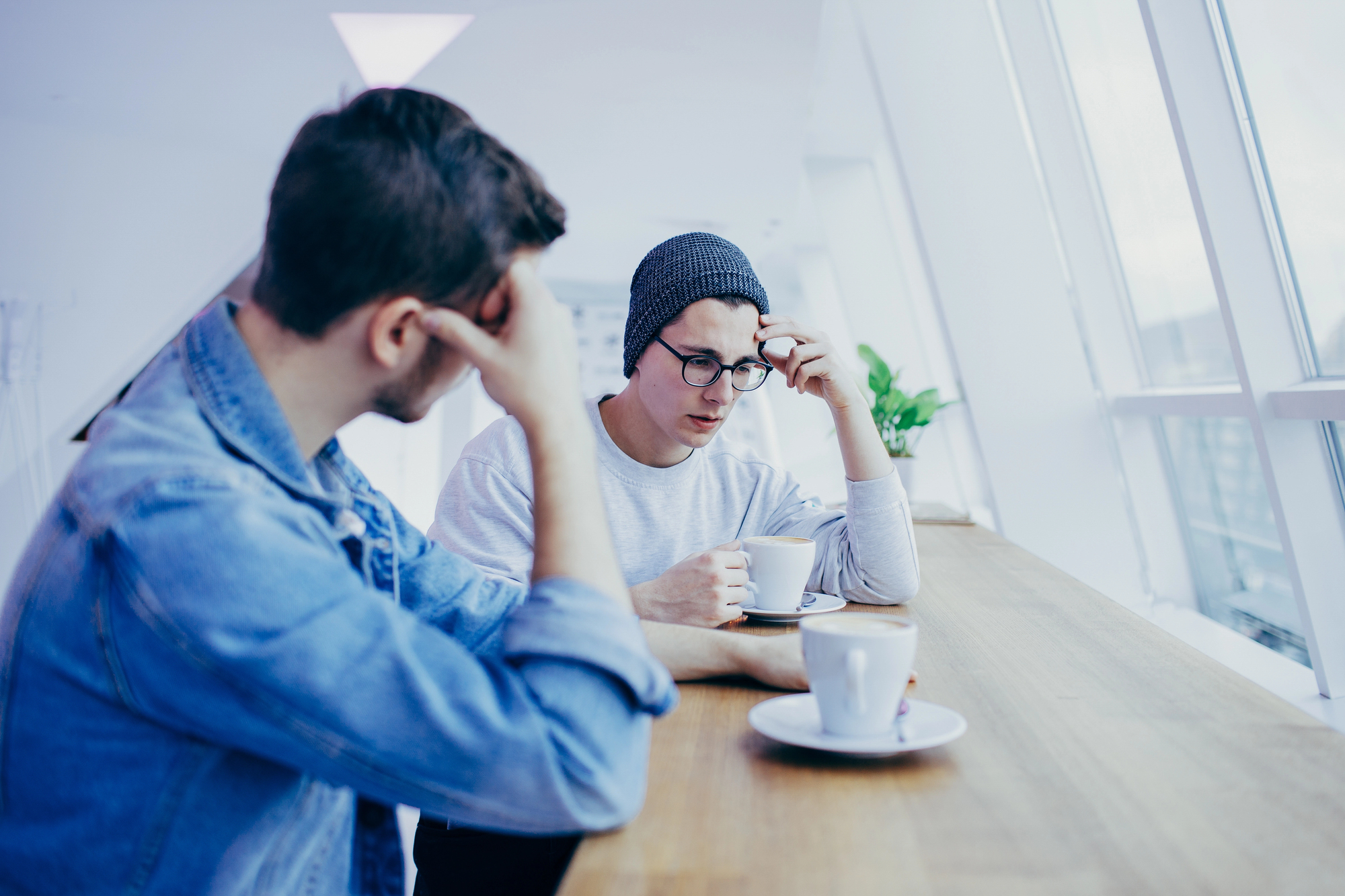 Two young men are seated at a wooden counter near a large window, each holding a coffee cup. One, wearing a beanie and glasses, looks thoughtful and engaged, while the other, in a denim jacket, rests his head in his hand, appearing contemplative.