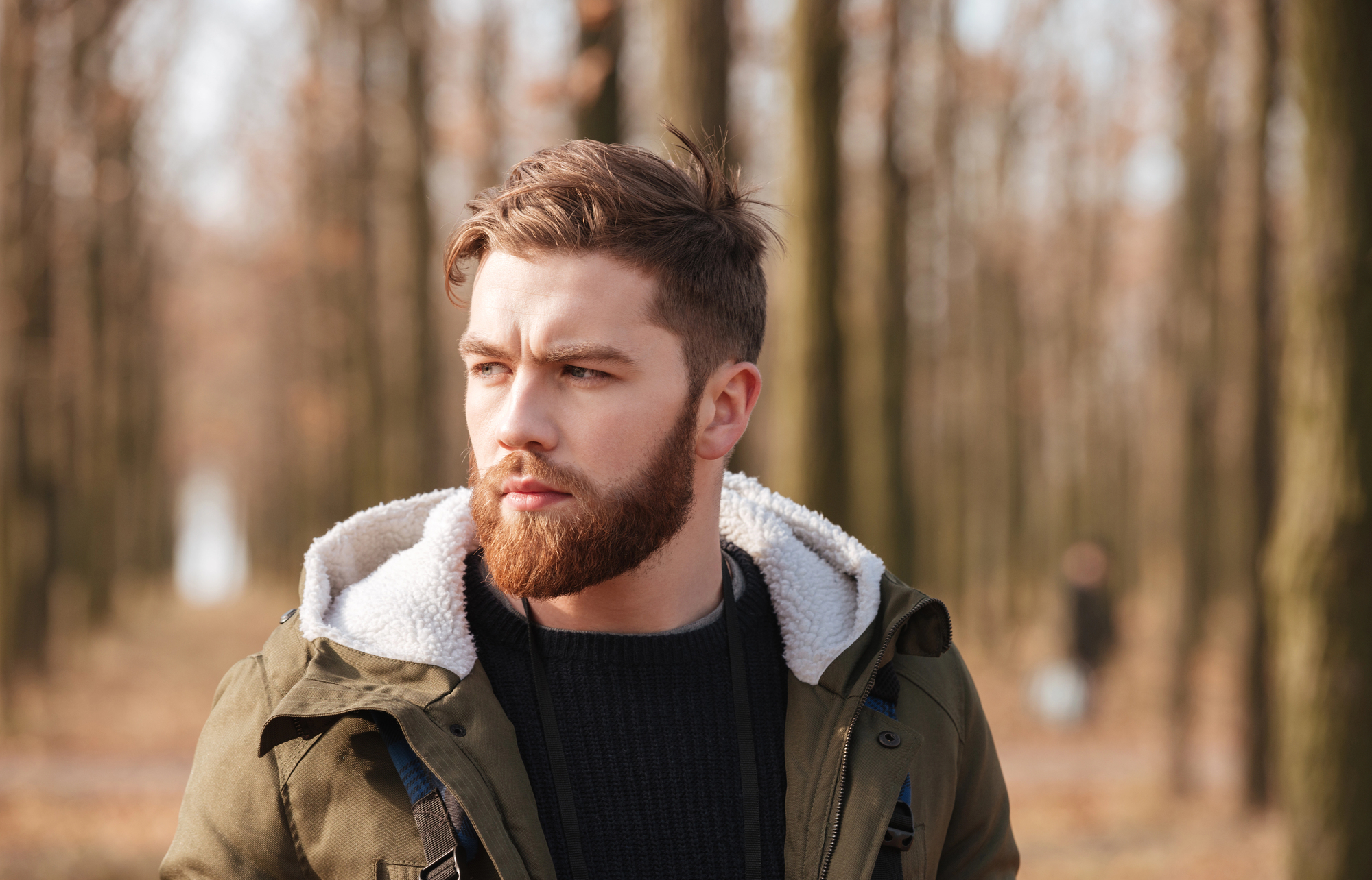 A man with a beard and short hair stands outdoors among tall trees, wearing a green jacket with a fleece-lined hood and a black sweater. The background is softly blurred, showing a path and more trees, with a person in the distance.