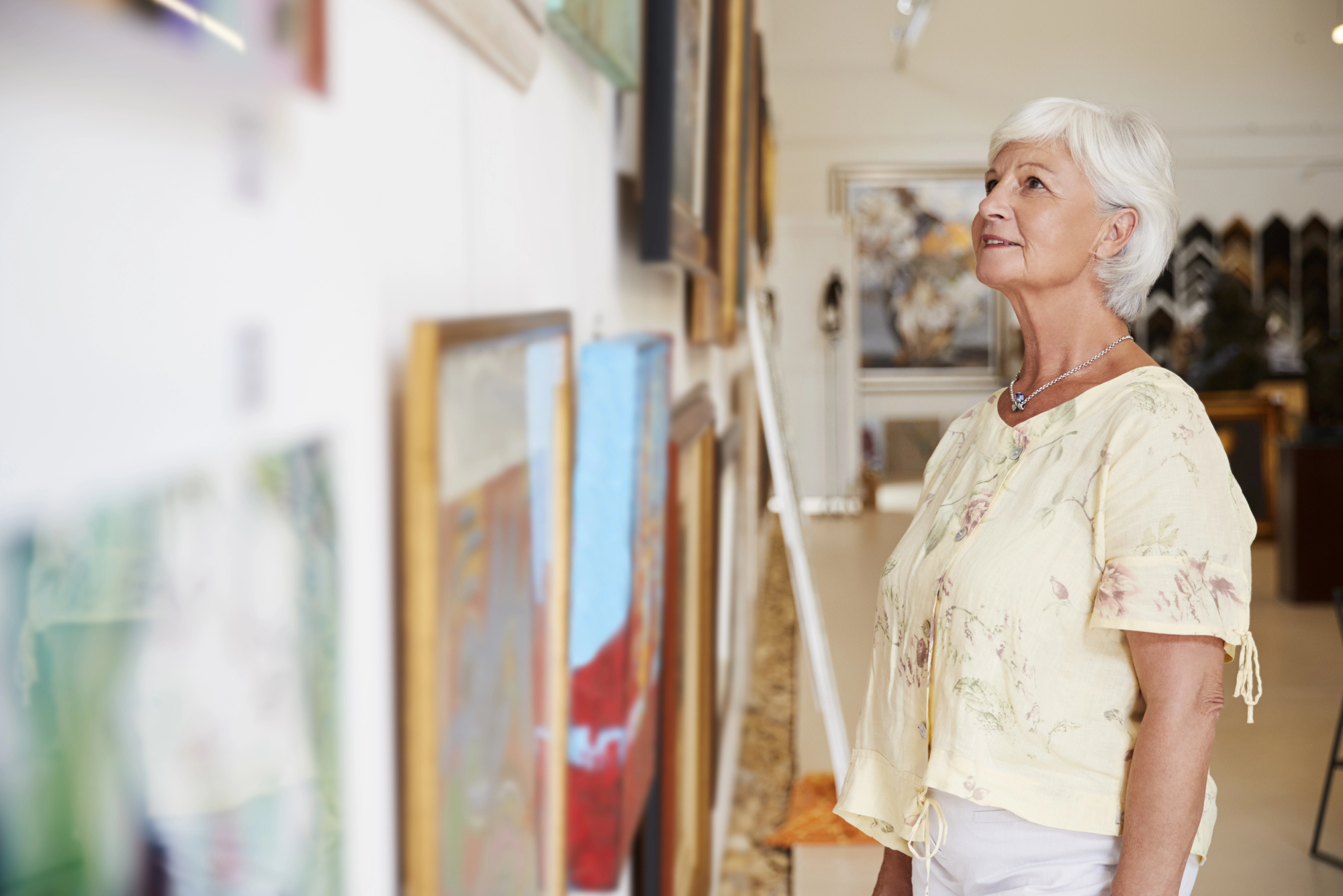 A woman with short white hair, wearing a light yellow blouse, stands in an art gallery looking thoughtfully at framed paintings on the wall. The background is softly focused, displaying more artwork.