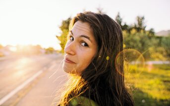 A woman with long hair looks back over her shoulder with a slight smile. She is standing outdoors on a sunny day, with a road and trees in the background. The sun creates a lens flare effect in the image.