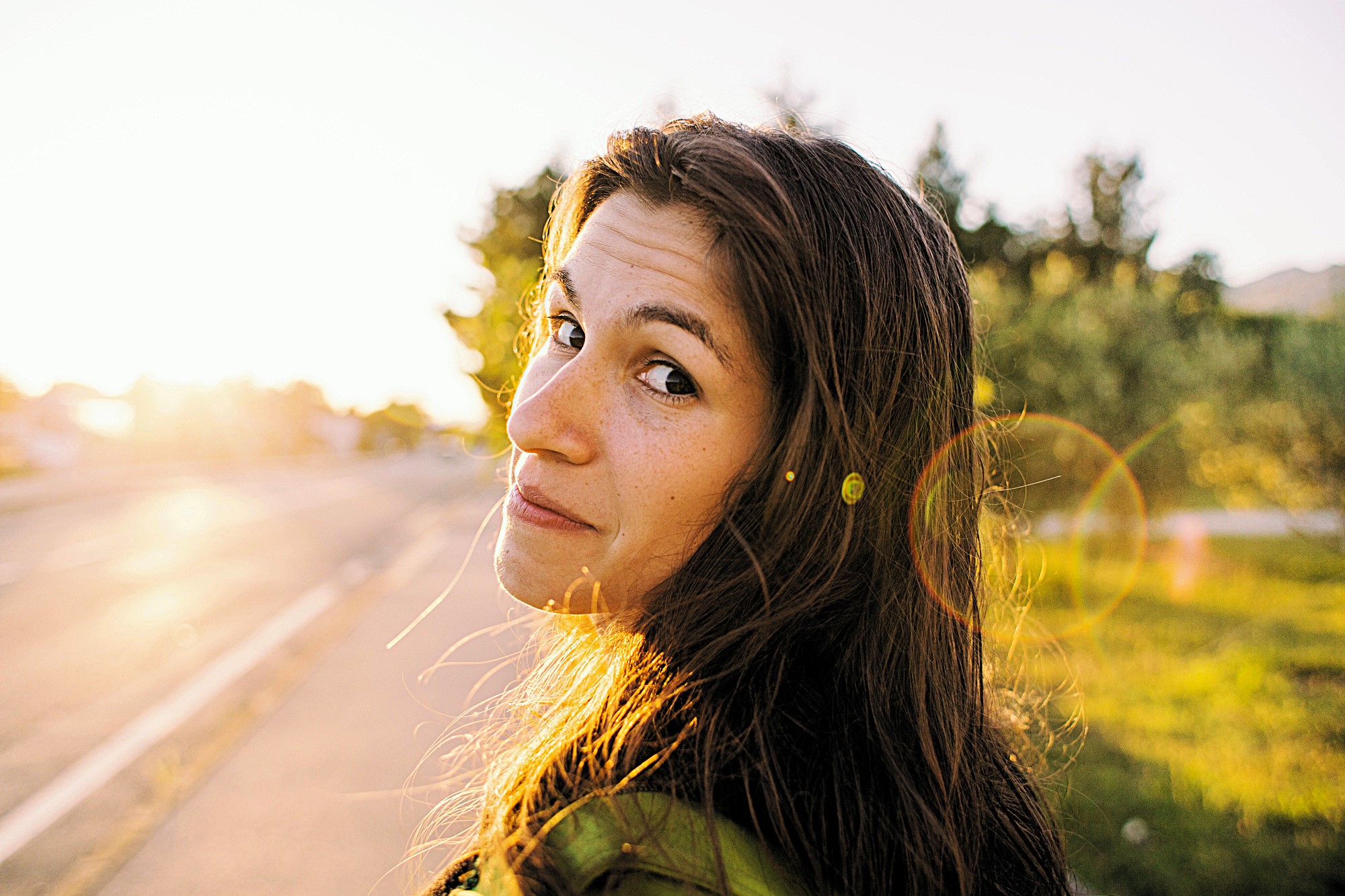 A woman with long hair looks back over her shoulder with a slight smile. She is standing outdoors on a sunny day, with a road and trees in the background. The sun creates a lens flare effect in the image.