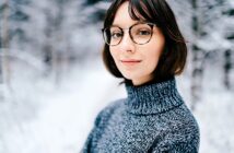 A person wearing glasses and a gray turtleneck sweater stands outdoors in a snowy forest, smiling softly. The background is blurred, featuring snow-covered trees.