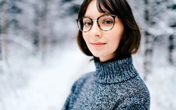 A person wearing glasses and a gray turtleneck sweater stands outdoors in a snowy forest, smiling softly. The background is blurred, featuring snow-covered trees.