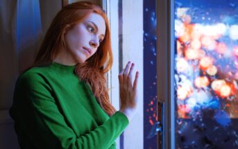 A young woman with long red hair, wearing a green sweater, leans against a window. She looks thoughtfully outside. The window is wet with raindrops, showing blurred colorful lights of a rainy evening cityscape in the background.