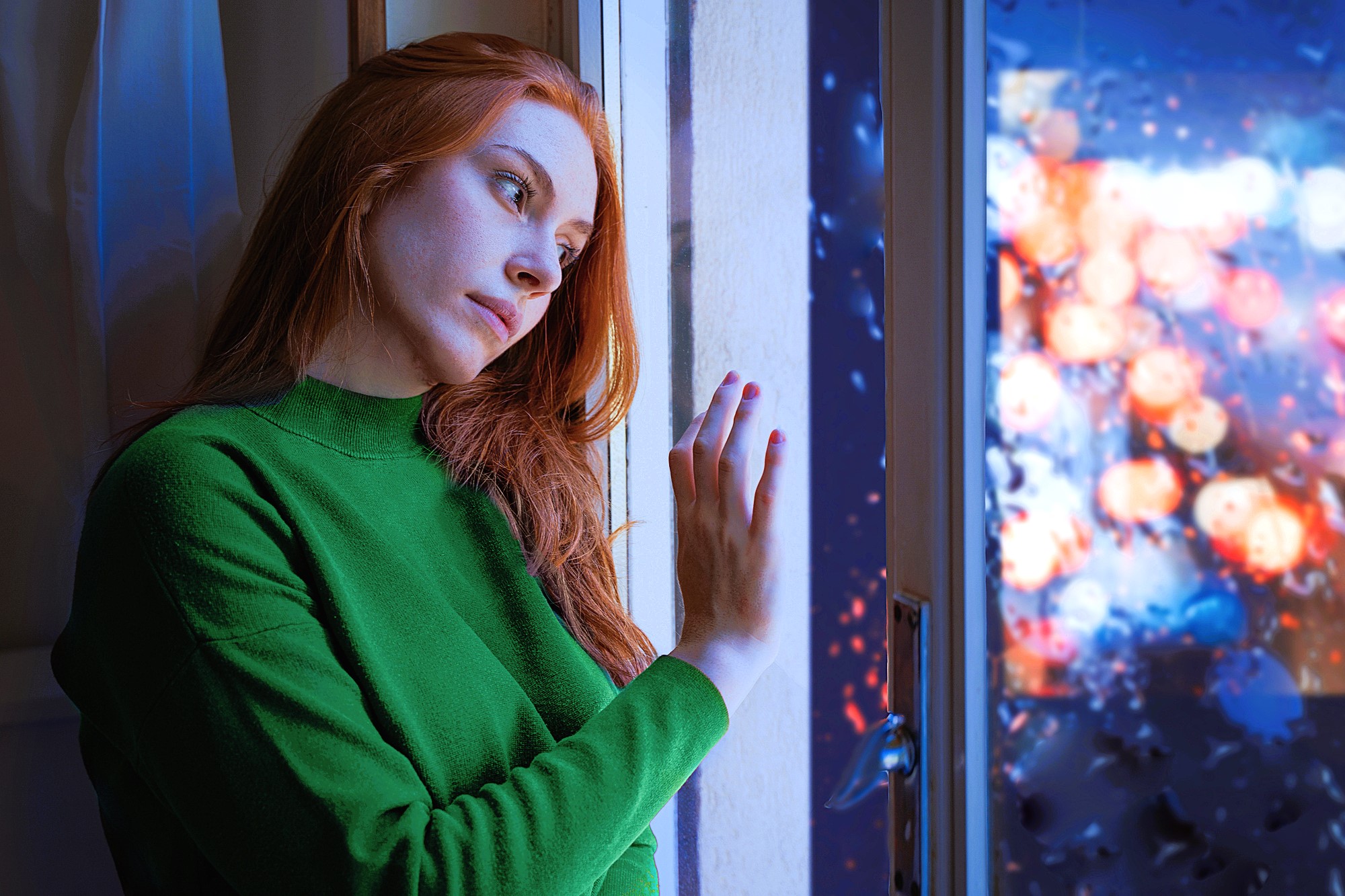 A young woman with long red hair, wearing a green sweater, leans against a window. She looks thoughtfully outside. The window is wet with raindrops, showing blurred colorful lights of a rainy evening cityscape in the background.