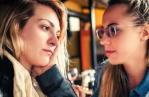 Two women sitting closely, engaging in a thoughtful conversation. One with long blonde hair rests her chin on her hand, while the other, wearing glasses, listens attentively. Background suggests a cozy indoor setting.