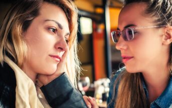 Two women sitting closely, engaging in a thoughtful conversation. One with long blonde hair rests her chin on her hand, while the other, wearing glasses, listens attentively. Background suggests a cozy indoor setting.