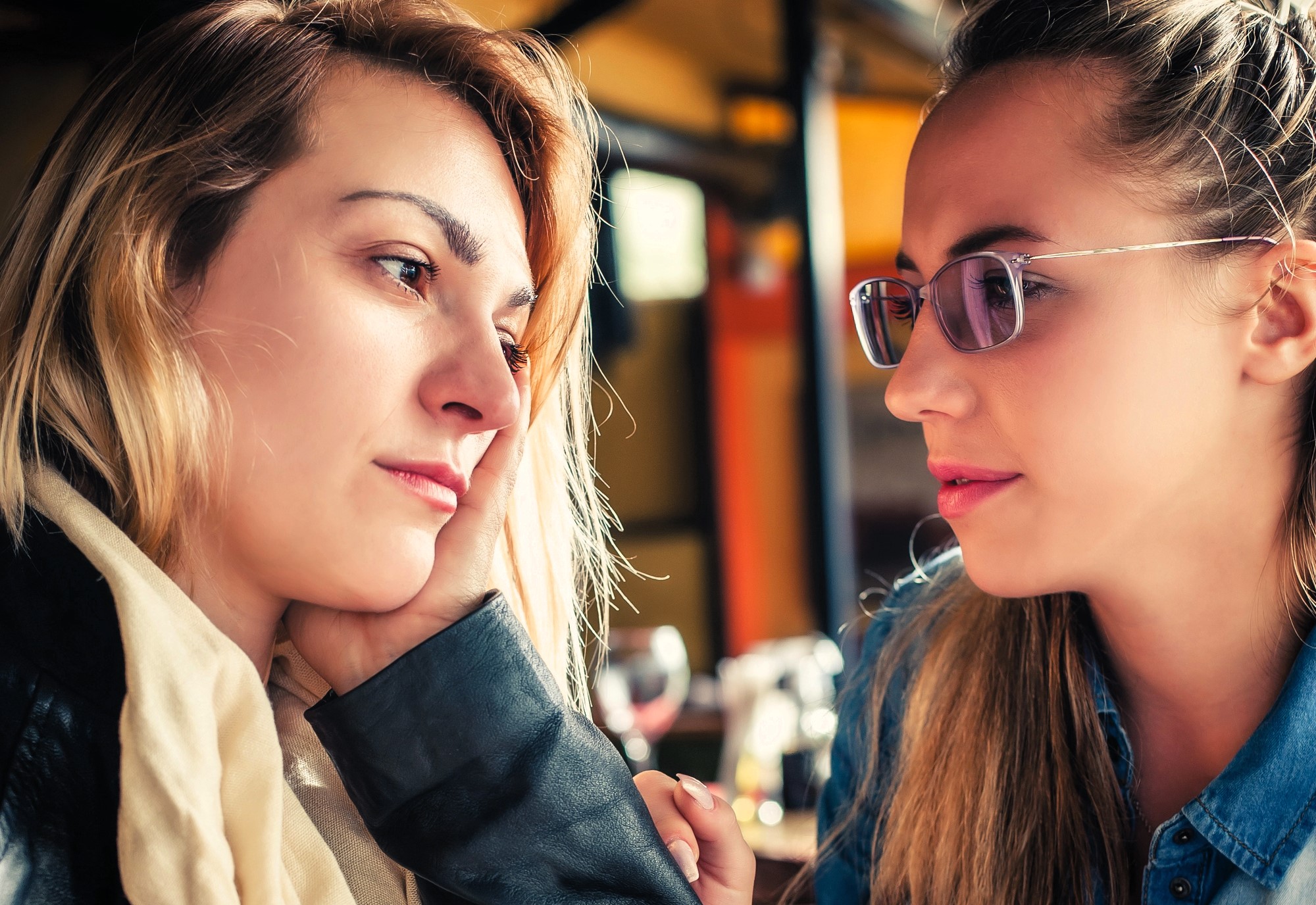 Two women sitting closely, engaging in a thoughtful conversation. One with long blonde hair rests her chin on her hand, while the other, wearing glasses, listens attentively. Background suggests a cozy indoor setting.