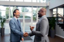 Two men in suits are shaking hands and smiling inside a modern office building. One has a beard and long hair, while the other is bald and wearing glasses. Large windows and greenery are visible in the background.