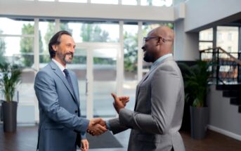 Two men in suits are shaking hands and smiling inside a modern office building. One has a beard and long hair, while the other is bald and wearing glasses. Large windows and greenery are visible in the background.