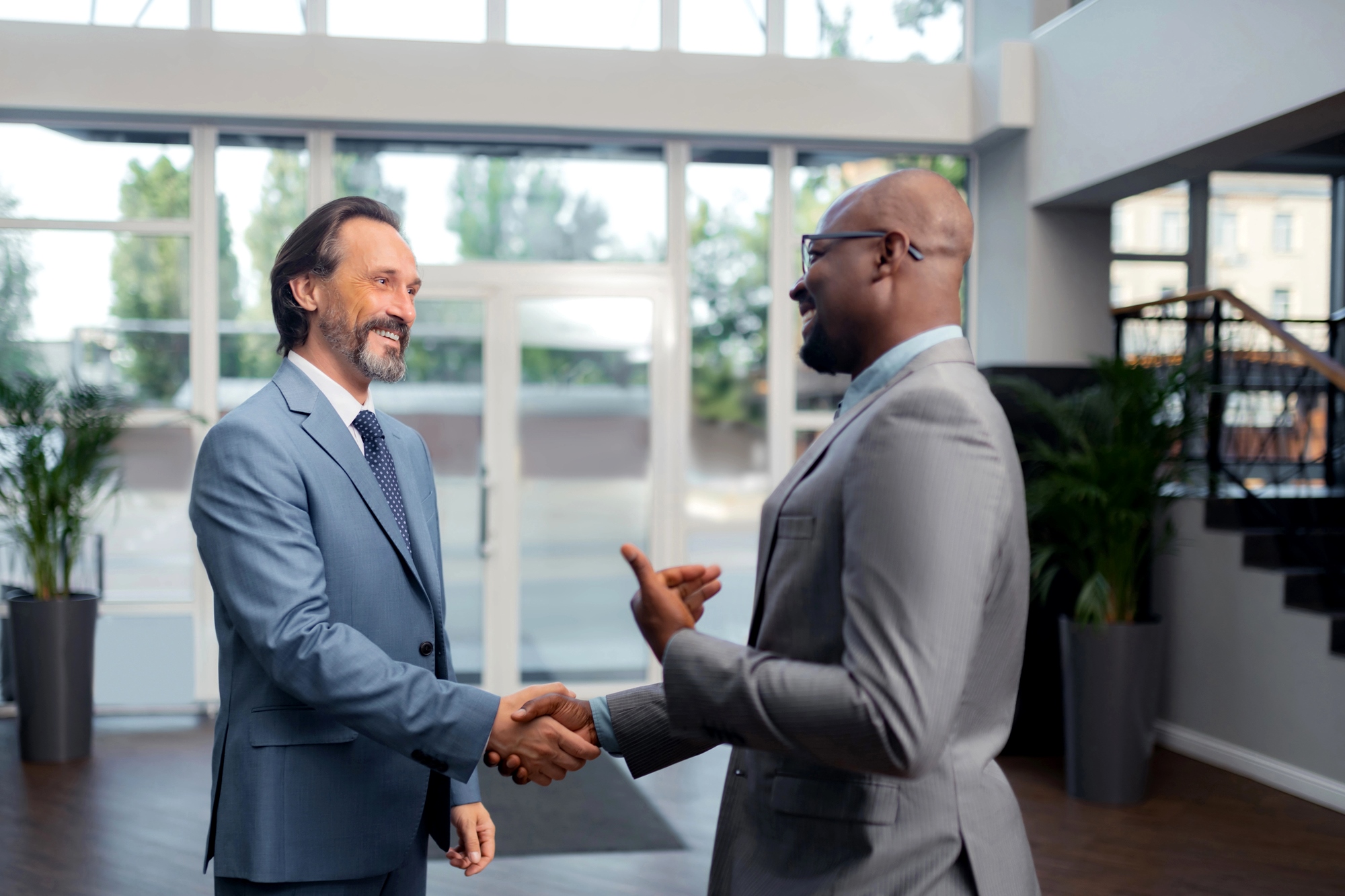 Two men in suits are shaking hands and smiling inside a modern office building. One has a beard and long hair, while the other is bald and wearing glasses. Large windows and greenery are visible in the background.