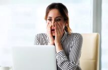 A woman in a striped shirt sits at a desk, looking at a laptop with a surprised expression. Her hands are on her cheeks, and she appears to be in an office setting.