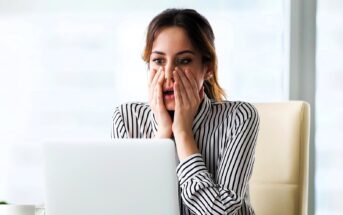 A woman in a striped shirt sits at a desk, looking at a laptop with a surprised expression. Her hands are on her cheeks, and she appears to be in an office setting.