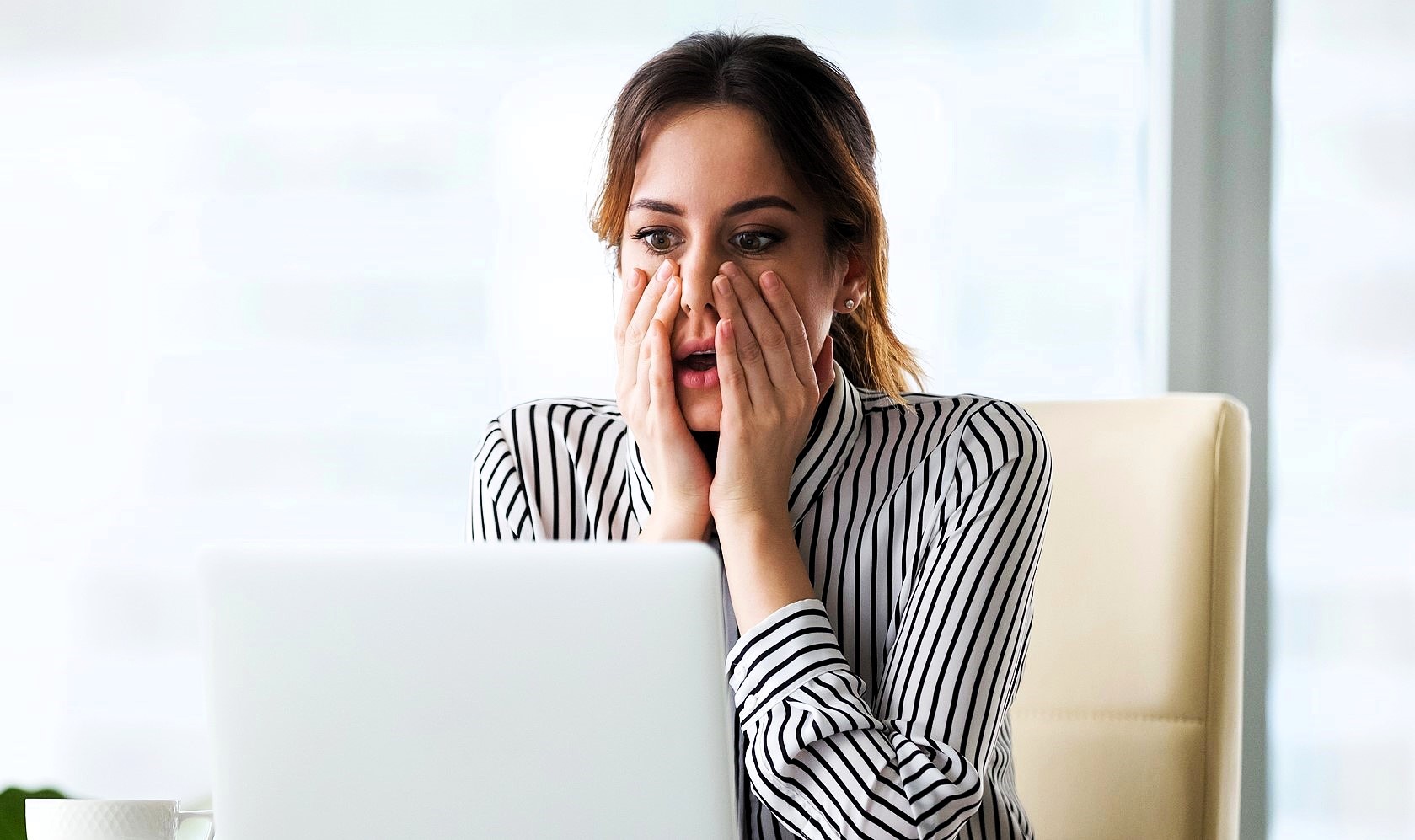 A woman in a striped shirt sits at a desk, looking at a laptop with a surprised expression. Her hands are on her cheeks, and she appears to be in an office setting.