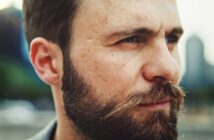 Close-up of a man with a beard and mustache looking to the side. The background is blurred, showcasing an outdoor environment with hints of greenery and building silhouettes. The man has a contemplative expression on his face.