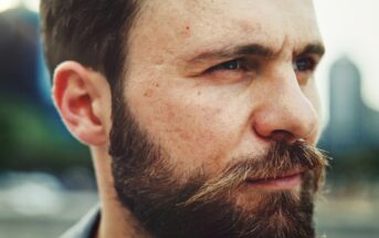 Close-up of a man with a beard and mustache looking to the side. The background is blurred, showcasing an outdoor environment with hints of greenery and building silhouettes. The man has a contemplative expression on his face.