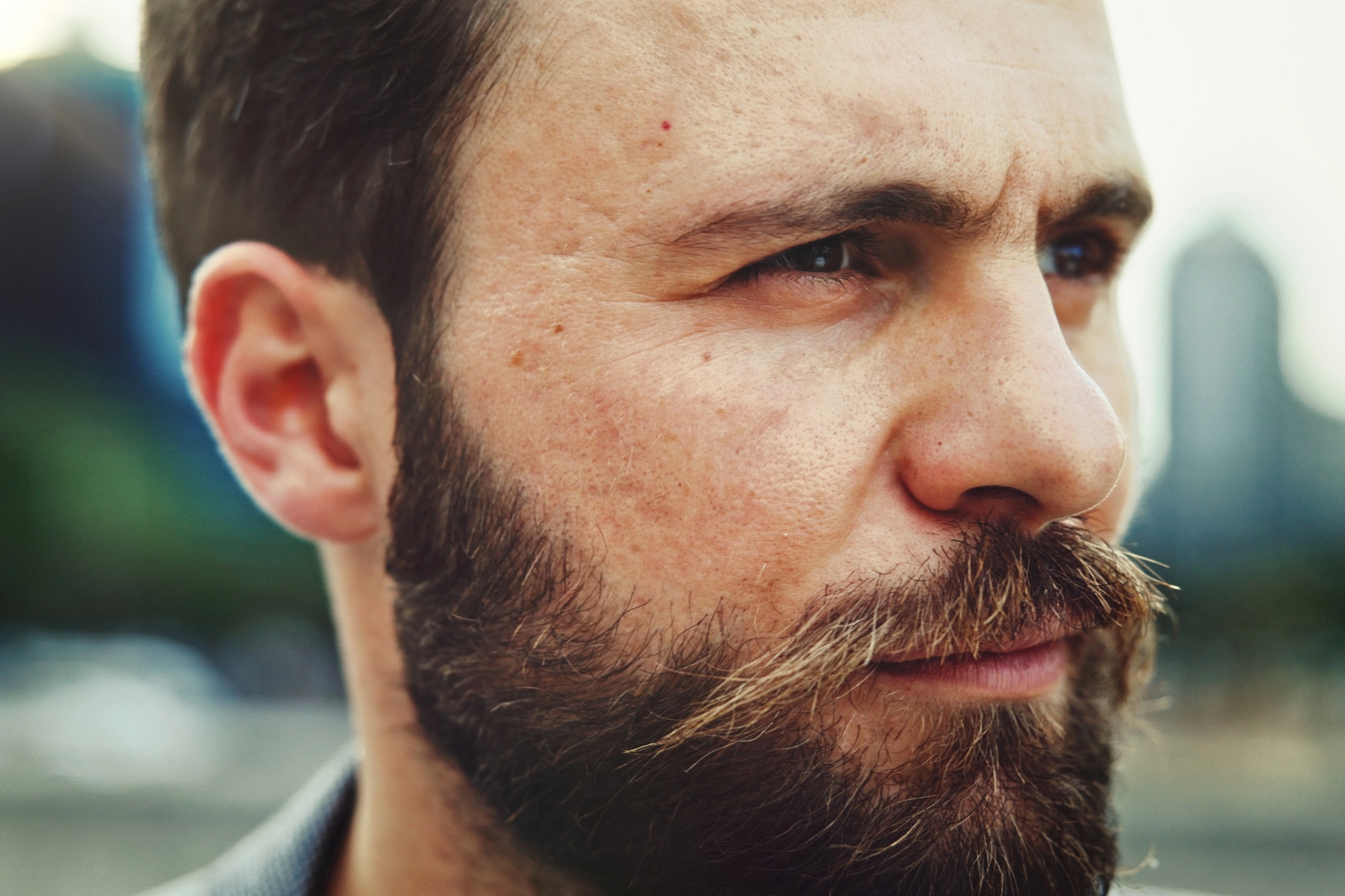 Close-up of a man with a beard and mustache looking to the side. The background is blurred, showcasing an outdoor environment with hints of greenery and building silhouettes. The man has a contemplative expression on his face.