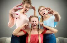 Three women in colorful tops acting playfully dramatic. Two women are leaning toward the third woman with exaggerated expressions, while the third covers her ears, pretending to ignore them. All are smiling, suggesting a fun, humorous moment.