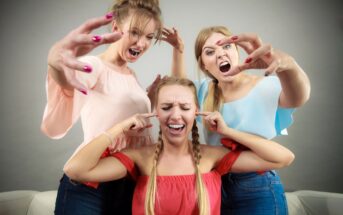 Three women in colorful tops acting playfully dramatic. Two women are leaning toward the third woman with exaggerated expressions, while the third covers her ears, pretending to ignore them. All are smiling, suggesting a fun, humorous moment.