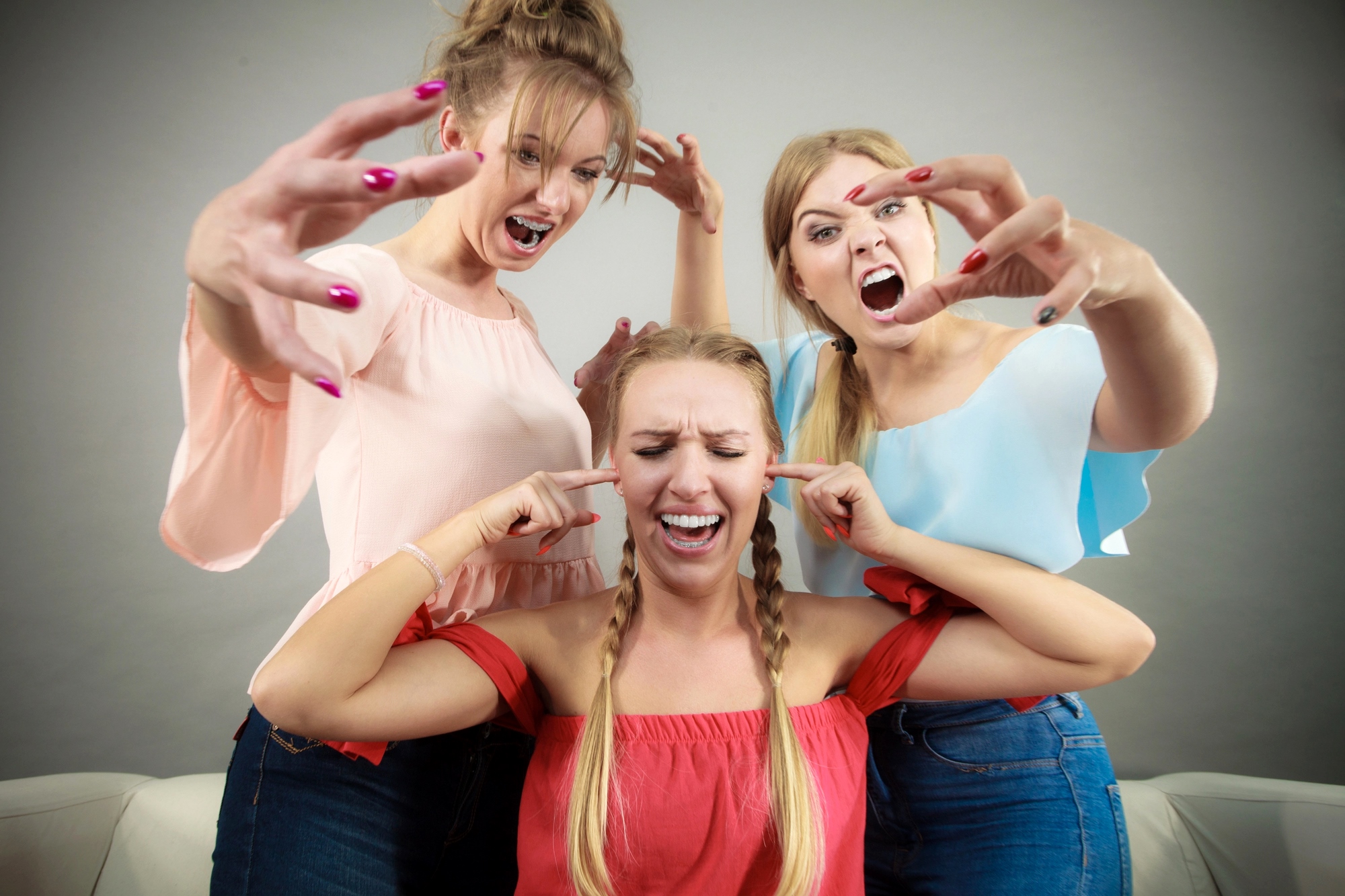 Three women in colorful tops acting playfully dramatic. Two women are leaning toward the third woman with exaggerated expressions, while the third covers her ears, pretending to ignore them. All are smiling, suggesting a fun, humorous moment.