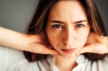 A person with long hair gazes intently at the camera, resting their hands on their neck. They wear a white shirt and have a neutral expression. The background is blurred.