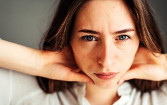 A person with long hair gazes intently at the camera, resting their hands on their neck. They wear a white shirt and have a neutral expression. The background is blurred.