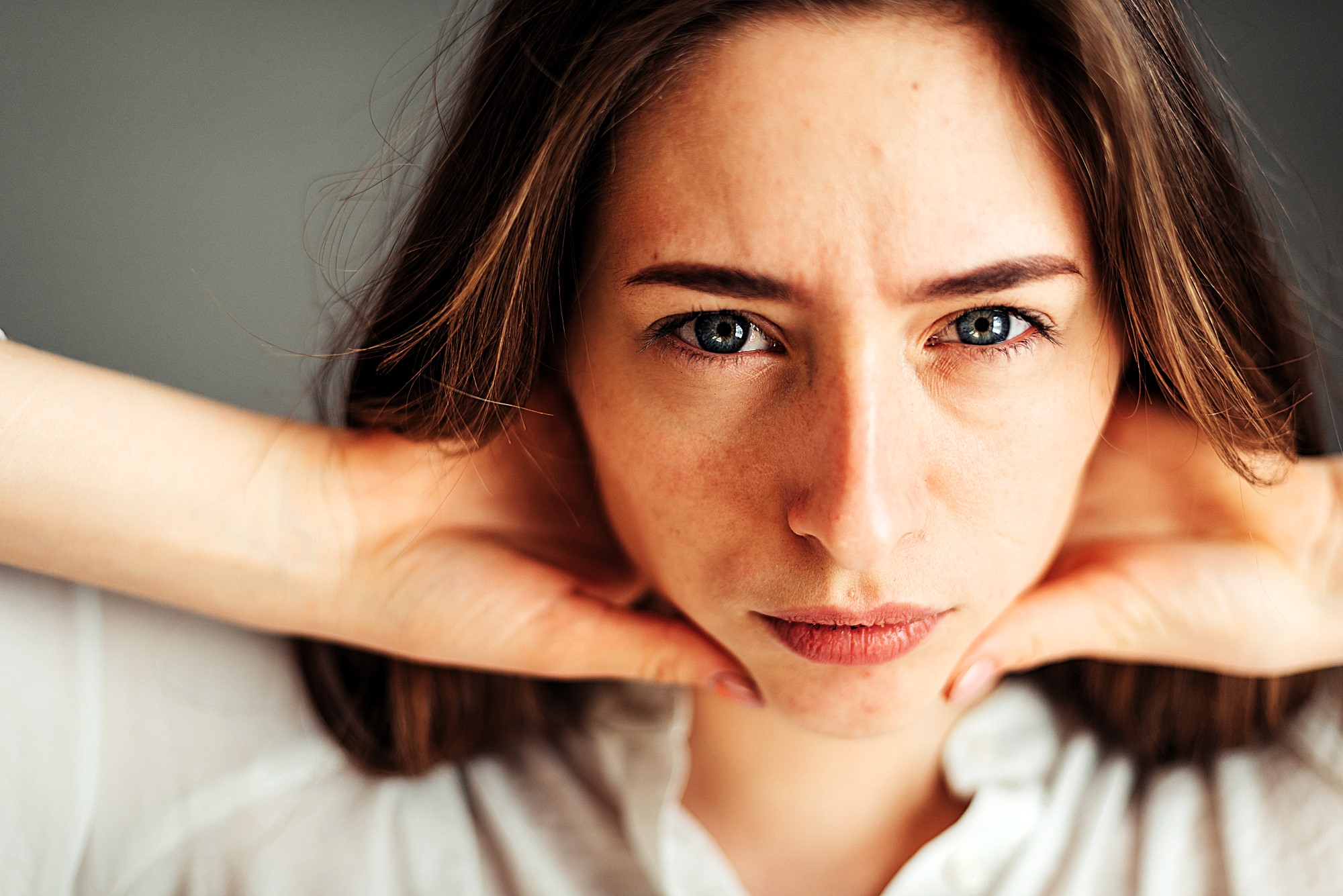 A person with long hair gazes intently at the camera, resting their hands on their neck. They wear a white shirt and have a neutral expression. The background is blurred.