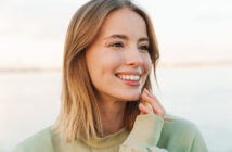 A woman with shoulder-length blonde hair smiles brightly, looking slightly to the side. She wears a light green sweater and touches her face gently with her hand. The background is softly blurred, suggesting an outdoor setting.