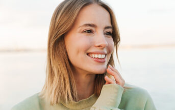 A woman with shoulder-length blonde hair smiles brightly, looking slightly to the side. She wears a light green sweater and touches her face gently with her hand. The background is softly blurred, suggesting an outdoor setting.