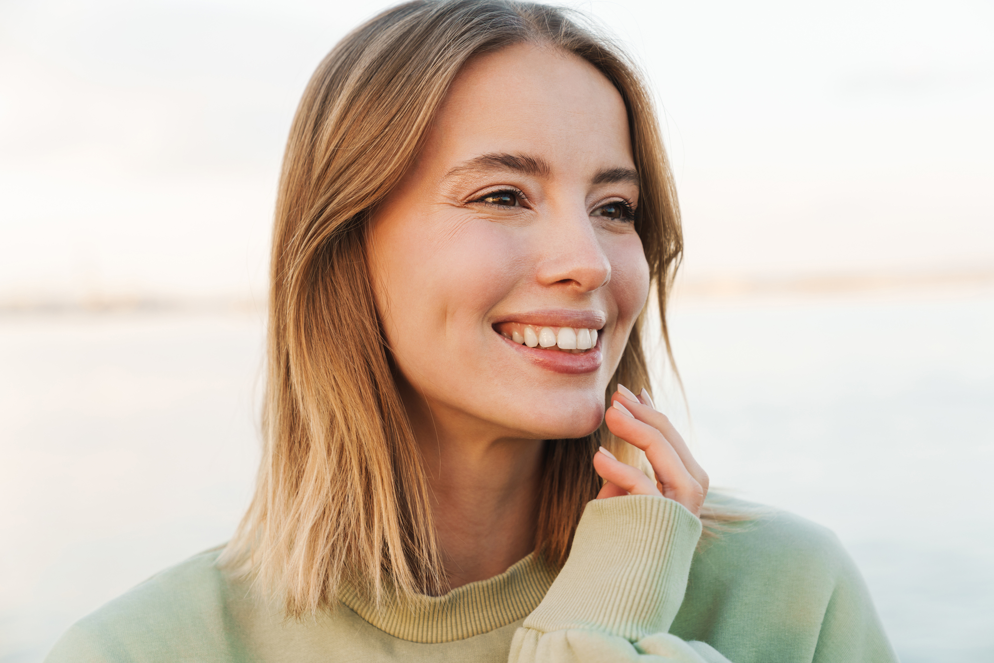 A woman with shoulder-length blonde hair smiles brightly, looking slightly to the side. She wears a light green sweater and touches her face gently with her hand. The background is softly blurred, suggesting an outdoor setting.