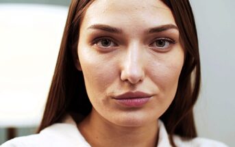 A woman with long brown hair looks directly at the camera with a neutral expression. She is wearing a white top, and the background is softly blurred, suggesting an indoor setting.