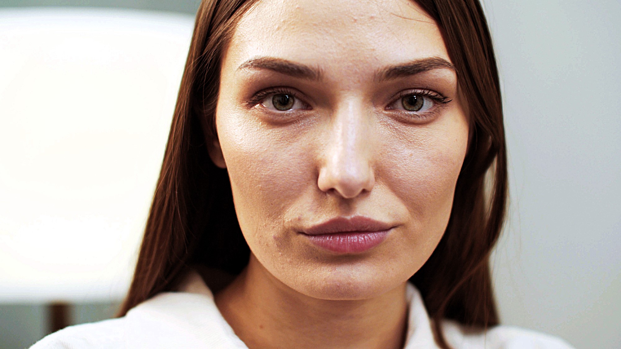 A woman with long brown hair looks directly at the camera with a neutral expression. She is wearing a white top, and the background is softly blurred, suggesting an indoor setting.